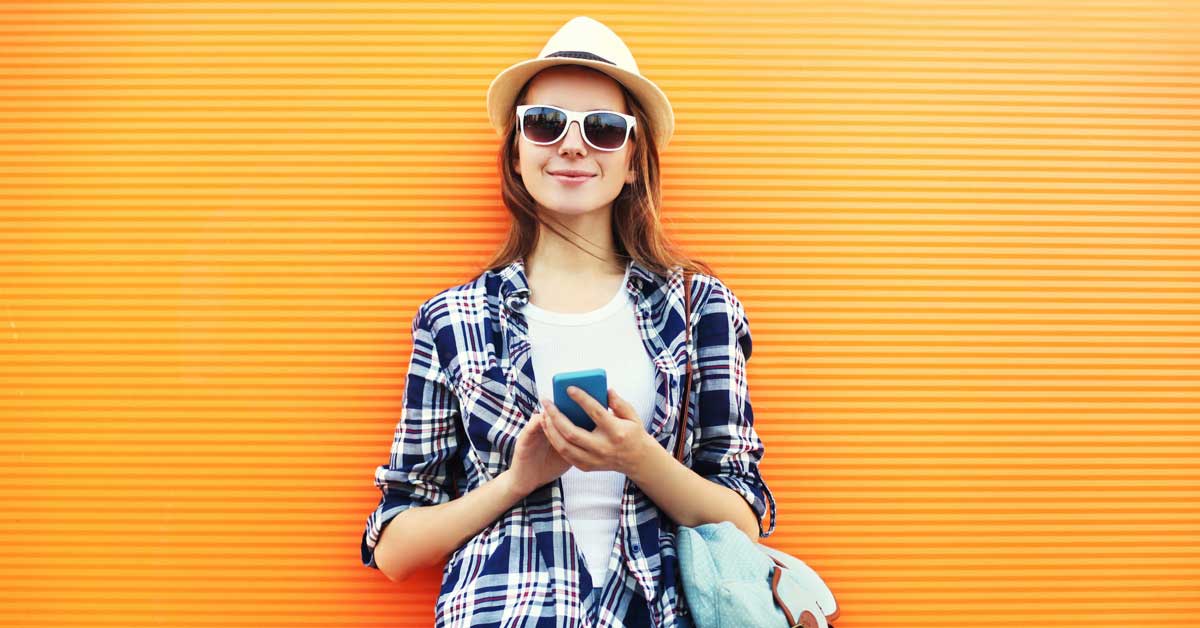 Woman standing in front of orange wall, smiling with sunglasses and a hat on, holding her mobile phone.