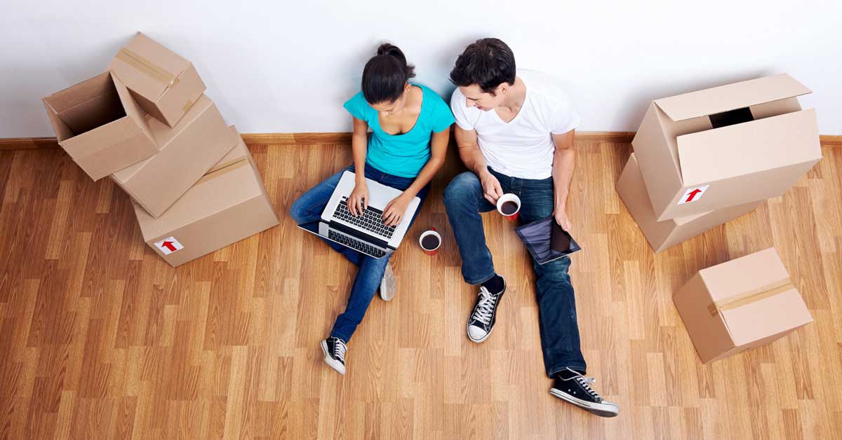 Aerial view from above of a man and woman sitting down with a laptop and drinking coffee, beside boxes that they are unpacking in their new home