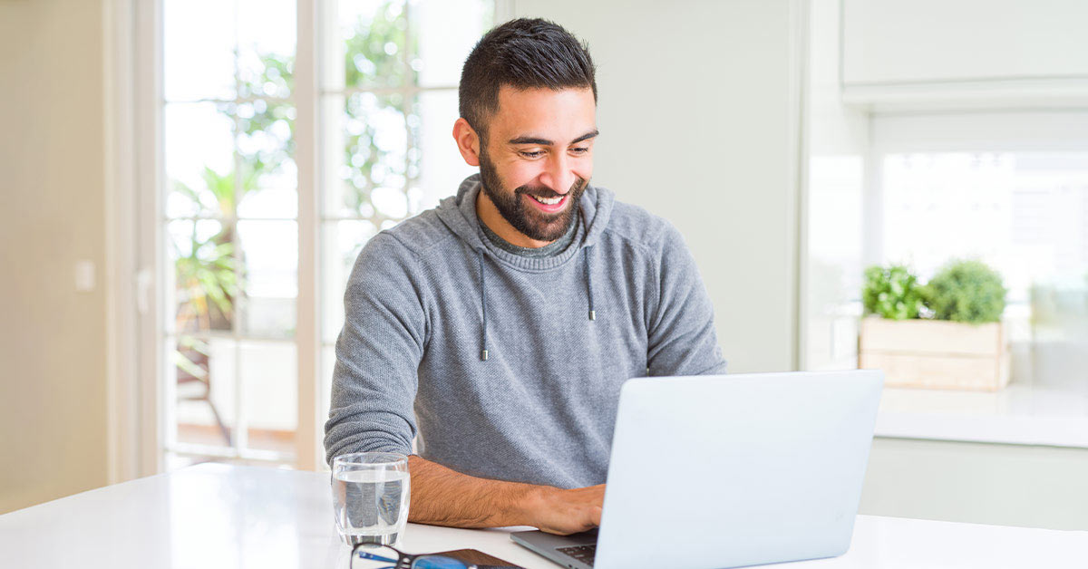 Man sitting at a kitchen counter using a laptop. He is smiling and there's a glass of water beside him.