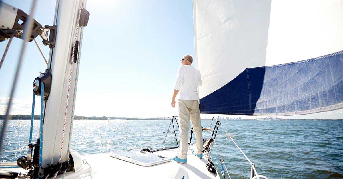 A middle aged men wearing a white top with sleeves rolled up and beige pants standing on a sail boat looking out towards the ocean with the main sail billowing in the wind.