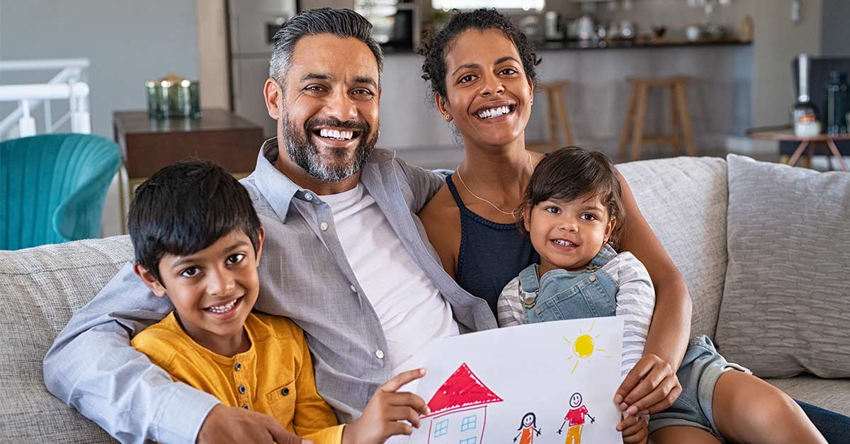 A family sitting on a couch smiling. There is mum, dad, and two children - a son and a daughter. They're holding a piece of children's art, which has a house and people drawn on it.