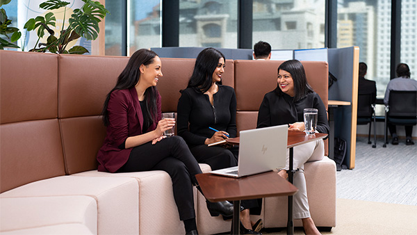 Three women sit on a couch. They're all smiling and talking. There is a small table in front of them with a laptop on it.