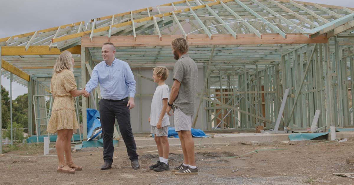 Two parents and their son stand in front of the frame of a new house, mother is shaking hands with a professional man
