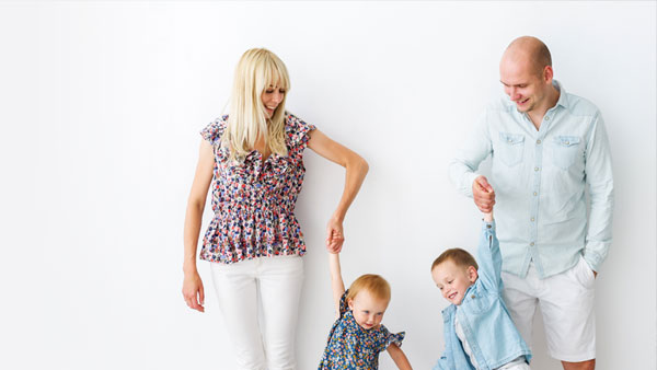 Mum and dad stand against a white wall with their two children between them. Each of them is holding one of their son's hands, and their sons are holding each other's hands.