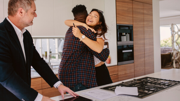 Close up of heterosexual couple sitting at desk with real estate agent, woman can be seen to be smiling. Real estate agent is handing the woman keys, and the keys are the focus of the image.