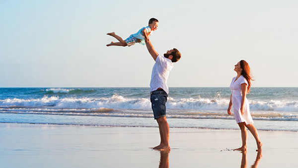 A couple standing on the beach with their young kid. The father is swinging the child happily into the air.