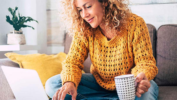 Woman sitting on a couch wearing a yellow sweater and holding a mug in one hand. She's using a laptop and has curly hair.
